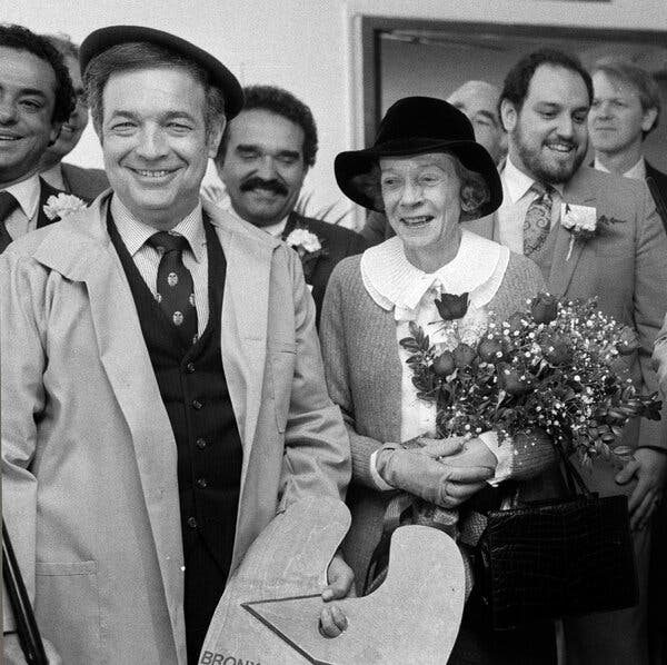 A black and white photo of a smiling Mr. Simon, wearing a dark beret, alongside Ms. Astor, who carries a bouquet of flowers. They are surrounded by other men wearing carnations on their lapels. 