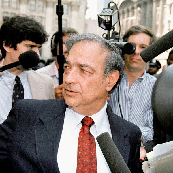A close-up photo of a man with gray, thinning hair and wearing a charcoal gray suit and red patterned tie as he is surrounded by broadcast newsmen with microphones.